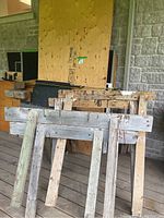 Two sets of hand crafted wooden saw horses made of weathered wood leaning against plywood sheets and other wood pieces on a wooden deck