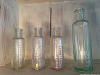 Four antique liniment bottles in varying glass colors (clear, light green, pink) with embossed lettering, standing vertically against a white wood background