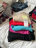 Stack of six folded cashmere sweaters in various colors with retail tags visible on beige sweater, lying on white quilted bedspread.