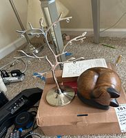 Photo showing a silver metal family tree-shaped frame with branches and loops, next to a carved wooden cat figurine on top of a cardboard box.