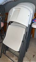 Stack of six white and grey Lifetime Products folding chairs folded and leaning against a wall.