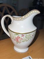 Full side view of a white ceramic cream pitcher with floral pattern and gold accents on wood table.