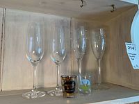 Four champagne flutes and two shot glasses displayed on a wooden shelf. Two plain flutes and two with Swarovski crystals can be seen along with two shot glasses, one black and one clear.