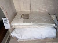 Two oblong table linens stacked; top one packaged Sophia damask tablecloth, bottom folded white cloth with textured checkered pattern.