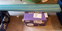 Rolled vinyl mat placed above purple box containing twin mattress on floor