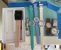 Photo showing all seven watches laid out on a blue surface, including the boxed smartwatch and six additional ladies watches with various strap types and colors.
