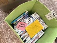 Top angled view of a green box filled with multiple white Wii game cases and some blue PS4 cases stacked inside, labeled as video games.