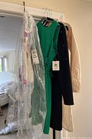 Four dresses and a blouse hanging on a rack, showing a floral dress in plastic cover, green sleeveless dress with tags, navy dress, and beige blouse.