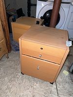 Two light wood tone particle board file cabinets with metal handles on drawers, one with visible lock hole, positioned on a floor near laundry appliances.