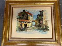 Framed oil painting showing a village street with buildings, archway, and floral accents. The painting is signed and in very good condition.