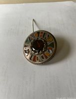 Scottish silver pin with round shape, central faceted brown stone, and multicolored enamel inlay surrounding it, skinny fastening needle visible.