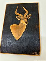 Front view of carved and painted wooden panel featuring antelope head artwork, showing signature in bottom right corner.