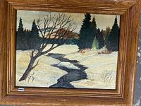 Framed oil painting showing a winter scene with snow, a flowing stream, and trees including one bare tree and several evergreen trees.