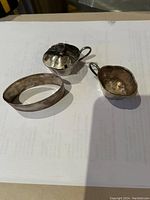 Photo showing three silver pieces including two small cups and one oval dish all placed on a white surface, tarnished and lightly used.