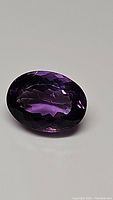 Close-up photo of a deep purple faceted oval amethyst gemstone on a plain background, showing the stone's cut and color.
