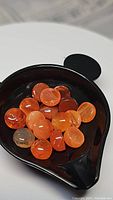 Photo of multiple small to medium round cabochon mandarin orange carnelian stones arranged in a black dish, showing their color, size, and polished smooth surface with some inclusions.