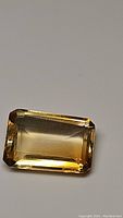 Close-up photo of a rectangular emerald cut golden citrine gemstone showing its transparency, cut facets, and color.