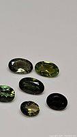 Six natural green oval cut tourmaline gemstones displayed, showing mixed shades of green and good clarity.