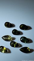 Eight pear-shaped green tourmaline gemstones arranged on white background, showing varying green shades and faceting details.
