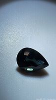 Pear-shaped natural sapphire with dark blue color and green undertone, shown on textured background with lighting to highlight facets.