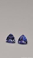 Pair of natural trillion cut tanzanite gemstones with strong purple-blue color on neutral background.