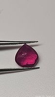 Close-up photo of ruby held by tweezers showing pear teardrop cut and rich purplish-red color with visible internal inclusions
