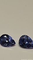 Close-up photo showing two pear-shaped natural tanzanite gemstones with deep purple-blue color placed side-by-side on a plain background.