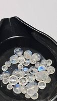 Parcel of round cabochon rainbow moonstone gemstones in black tray, showing blue flash and inclusions under white lighting.