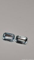 Two emerald cut sky blue topaz gemstones laid side by side on a plain surface, showing clarity and color.