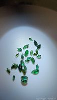 Photo showing a scattered group of small marquis cut emerald gemstones on a white background with spotlighting highlighting their green color and facets.