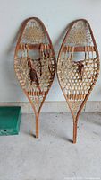 Pair of antique wooden snowshoes with woven rawhide webbing, shown standing upright against a white wall on a cement floor.