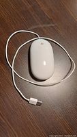 Top view photo of white Apple wired mouse with attached cable and USB connector on brown wood surface.