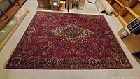 Full view of large rectangular area rug with red background, intricate floral and medallion patterns, and detailed border, placed on basement floor surrounded by boxes and shelves.