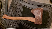 A curved wooden handled axe with a metal head covered by a brown leather sheath marked 'JACOBS HENCKEL'. The axe is placed on a dark textured background.