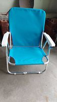 Blue folding beach chair with white armrests and silver aluminum frame, photographed indoors on a concrete floor.