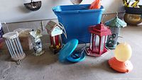 Image showing various bird feeders arranged on the ground including metal lantern-style, ceramic bird-shaped feeder, blue plastic feeder, and others.