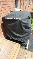 Broil King grill covered with a black protective cover showing the Broil King logo, positioned on a wooden deck next to a brick wall.