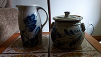 Photo showing a tall ceramic jug with handle and spout alongside a round ceramic pot with lid. Both pieces feature blue floral and leaf decorations on beige-gray ceramic.