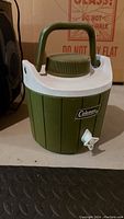 Green Coleman cooler with white top and spout placed on floor next to other items.