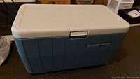 Photo showing side of cooler with blue ribbed body and white lid, Coleman Mariner label visible.