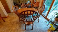 Oval wooden dining table with four matching spindle back chairs placed around it in a tile floor room with large windows.