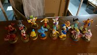 Photo of 14 Disney character figurines on round bases arranged in two rows on a wooden surface.