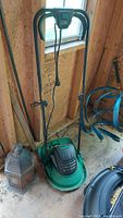 Photo showing an electric garden tool with a green base and black handle grips, a power cord hanging down. Positioned indoors against a wooden wall with window light.