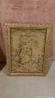 Photo showing embroidered art of a woman and man under tree, framed in ornate gold frame