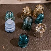 Eight vintage glass lightning rod insulators placed on a black rubber mat. Mix of clear, amber, blue, aquamarine colors and various shapes shown clearly.