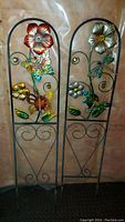 Two vertical metal lawn decorations featuring colorful 3D flowers and butterflies on black metal frames with curled vine and leaf designs.