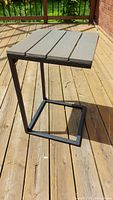 Metal side table with square black metal framework and four wooden slats painted grey on top, photographed outdoors on a wooden deck in sunlight.