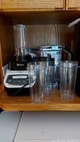 Ninja blender base with multiple clear plastic blending containers of various sizes and some lids, displayed on shelf.