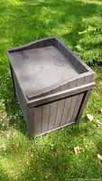 Closed dark brown plastic outdoor storage bin on grass, showing wood-like texture on panels and flat squared top lid with slight water marks.