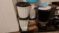 Photo showing eight round plastic storage bins stacked against a wall, five are white with white lids, three are black with black lids. Bins have ribbed vertical sides. One cardboard box is also visible.